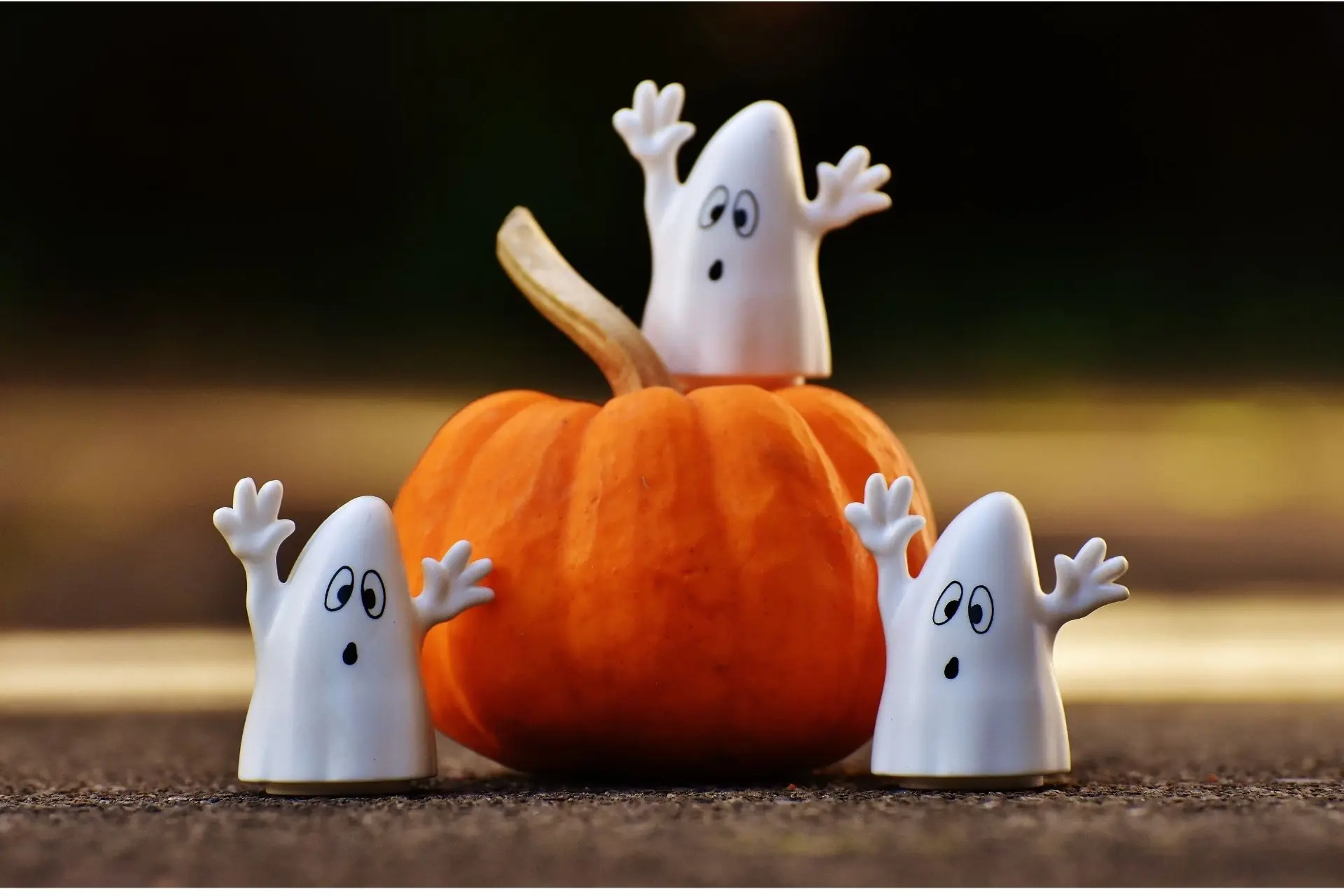 Three white cartoon ghost decorations surrounding an orange pumpkin, with one ghost perched on top and two standing beside it.